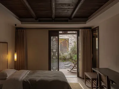 A serene minimalist bedroom with wooden and fabric elements, featuring a neatly made bed and a view of a serene outdoor area.