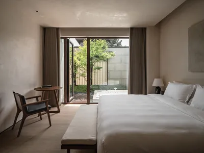 A minimalist bedroom with a white fabric bed on a wooden floor, featuring natural light and greenery outside through large wi