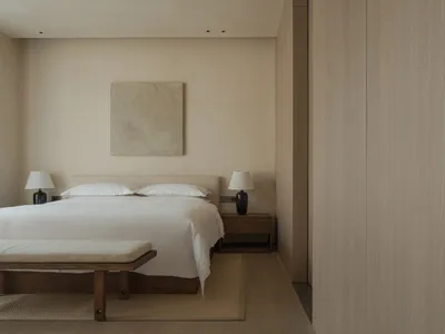 A minimalist bedroom with a bed, nightstands, and a bench, all in neutral colors and featuring a wooden headboard and a large