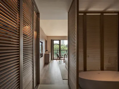 A minimalistic bedroom featuring wooden furniture and a freestanding bathtub, with a view of the outdoor balcony and greenery