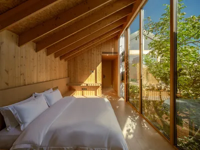 A minimalist bedroom with wooden walls and floor, featuring a comfortable bed with white bedding and a large window overlooki