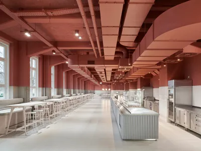 A modern restaurant interior with a concrete ceiling, long dining tables, and stainless steel counters.