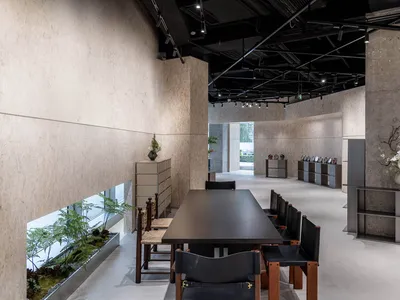 A modern restaurant interior featuring concrete walls, wooden furniture, a long dining table with black chairs, and plants.