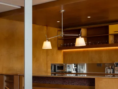 A modern kitchen with wooden cabinets, shelves, and stools, featuring a stainless steel countertop and warming lights.