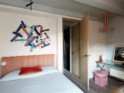 A minimalist bedroom with a pink bed, white bedding, and a colorful abstract sculpture on the wall.