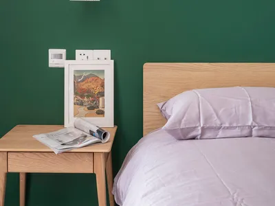 A minimalist bedroom with a wooden headboard on a green wall, featuring a bed with white bedding and a side table with a fram