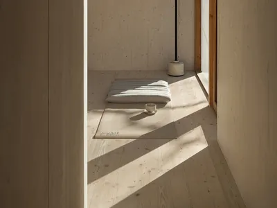Minimalist bedroom with wooden flooring and walls, a white bed, and sunlight streaming through the window.