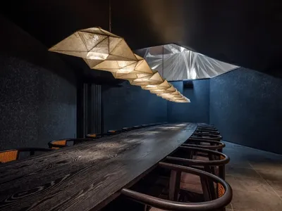 Long black wooden dining table with curved wooden chairs and geometric paper lampshades in a restaurant interior with dark blue walls and a skylight.