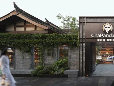 A man walks in front of an Asian restaurant with a black facade, featuring dining tables and chairs inside, creating an inviting and comfortable atmosphere
