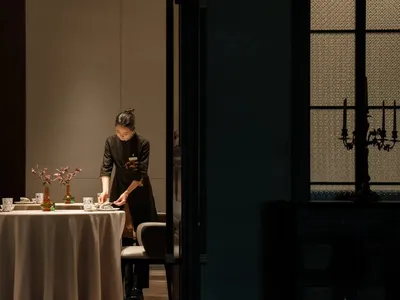 一位穿著圍裙的女性在專業廚房中於桌上準備食物，周圍有碗、湯匙和刀子