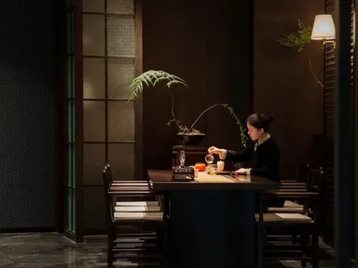 一位女性坐在餐桌旁，置身於燈光昏暗的房間，周圍環繞著椅子，營造出溫馨親密的氛圍