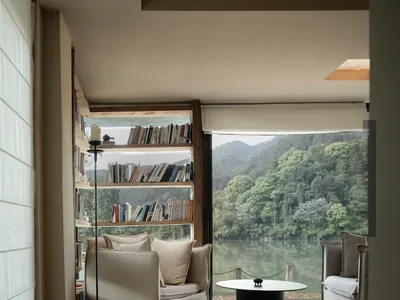 A bright and inviting living space with a large window overlooking a lake, featuring two couches, a chair, and a coffee table, surrounded by books and filled with natural light