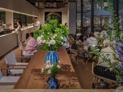 現代餐廳空間，木質地板、牆上藝術品、窗戶自然光線，大型餐桌與小型餐桌，人們入座交談