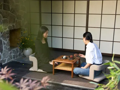 Two women sit in an Asian-style living area with a wooden table, surrounded by decorative vases and potted plants, creating a cozy atmosphere
