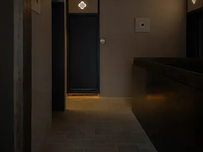 A dimly lit hallway with a black door, two clocks on the wall, and a remote control on the floor, creating a mysterious atmosphere