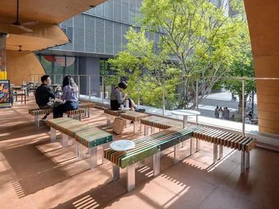 Dining area with wooden benches and tables, natural light from large windows, and several people enjoying meals in a warm and welcoming atmosphere