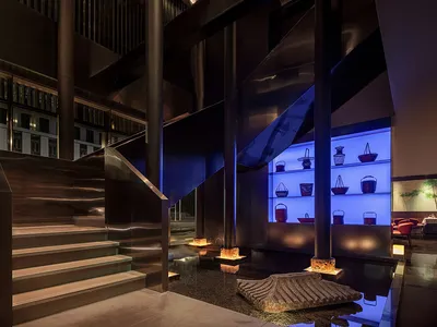 A modern hotel lobby with a wooden staircase, two chandeliers, and a warm atmosphere