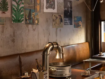 Casual restaurant interior with concrete walls, a bar, and dining table, featuring chairs and scattered bottles