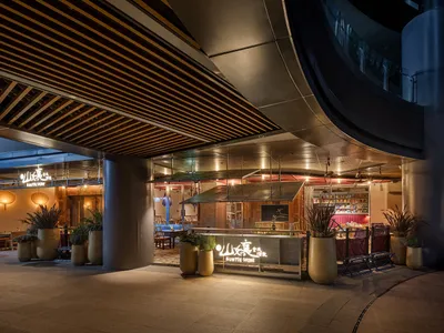 A modern Asian restaurant with a large dining area, tables, chairs, potted plants, artwork on the walls, and warm lighting