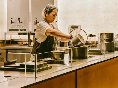 一位女性在工業風廚房中於檯面上準備食物，周圍環繞著碗盤與廚具，在注重細節的專業環境中