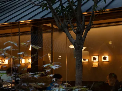 A casual dining area with people eating at tables, surrounded by chairs, artworks on the walls, and a mix of natural and artificial lighting