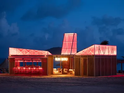 Coastal restaurant with red accents, pink lights, and chairs near the water's edge at night