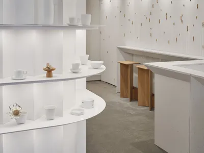 A well-lit kitchen showroom with white surfaces, a sink, countertop, and decorative items such as cups, bowls, vases, and potted plants