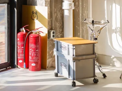 A cluttered living area with a window, red fire extinguishers, bottles, and a bowl, suggesting a functional yet somewhat disorganized space