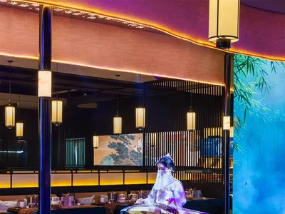 一位女士在溫馨亞洲餐廳中一張桌子前享用餐點，桌前有一幅大型畫作，周圍有多張桌椅、桌面擺放杯具，營造輕鬆氛圍