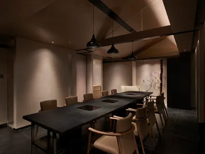Empty restaurant with a long wooden dining table surrounded by leather chairs and potted plants, creating a cozy atmosphere