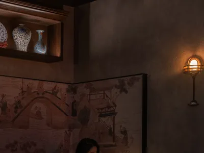 A woman sits at a dining table in an Asian restaurant with oriental artwork on the wall, surrounded by chairs and a comfortable atmosphere