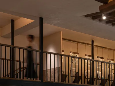 A calm and inviting interior space with a wooden staircase, stone elements, and soft diffused lighting, featuring visitors exploring the exhibits