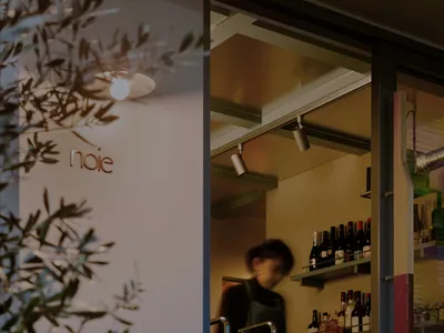 A cozy wine shop with a woman behind the counter, surrounded by bottles and chairs, creating a warm and inviting atmosphere