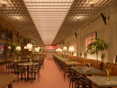 A modern restaurant interior with a long dining table, pink carpet, and potted plants, featuring wine glasses and a well-lit atmosphere