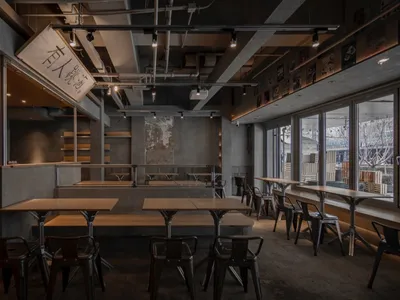 Empty industrial restaurant interior with a large dining table and multiple chairs, a couch in the background, and a commercial atmosphere