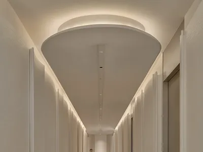 A long, modern hallway with white walls and floors, featuring several doors and a person standing in an adjacent room