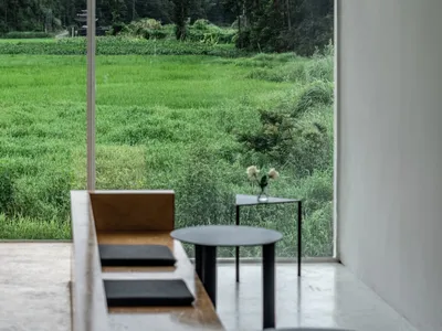 A calm restaurant interior with a dining table, chairs, couch, and vase, surrounded by a large window with a view of a green field, bathed in natural light