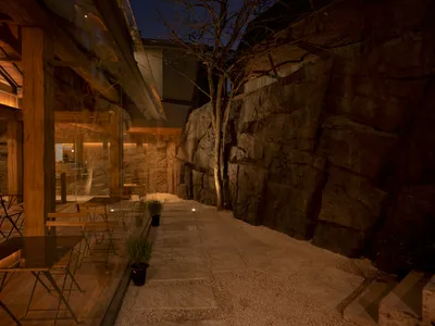 Outdoor dining area at night with tables, chairs, and potted plants in front of a lit building on a gravel pathway with a stone wall