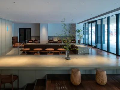 A restaurant dining area with multiple tables, chairs, and wood flooring, set against a backdrop of white and grey walls
