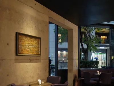 A modern restaurant with a large dining table and several smaller tables, purple accents, and a concrete finish, lit by lamps creating a warm ambiance