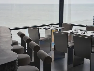 Sophisticated dining area with a large glass window offering a stunning ocean view, surrounded by chairs, a dining table set with wine glasses, cups, cutlery, and bottles