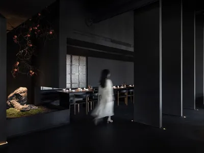 一位女子走過燈光昏暗的餐廳，內有餐桌、椅子，以及散置的杯碗