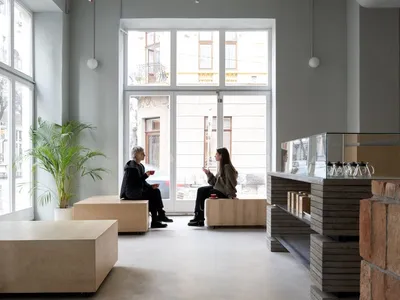 Modern living space with wooden floors, white walls, and large windows, featuring a cozy atmosphere with potted plants and two people sitting on couches