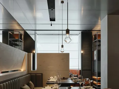 A dimly lit restaurant interior with modern furniture and table settings, featuring wine glasses, cups, and vases, creating a sophisticated atmosphere