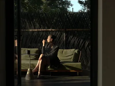 A woman sits on a couch in a calm and relaxing indoor living space with a dining table, chairs, and a vase in the background, near a window with natural light