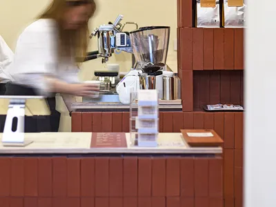 A barista prepares drinks at a busy coffee shop counter with various items and customers in the background