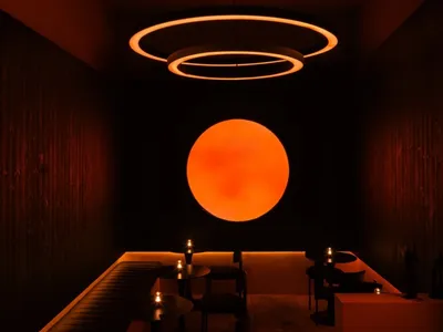 一間燈光昏暗的餐廳，內有兩張餐桌、椅子和花瓶，在黑色空間中被橘色太陽照亮