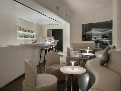 A modern restaurant bar with a counter and chairs, featuring white walls and flooring, and bottles on the counter