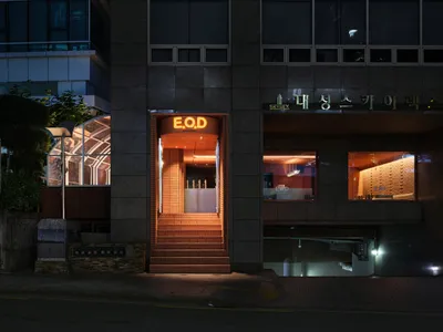 A modern Asian restaurant at night with a brick exterior, glass windows, and a warm interior atmosphere, featuring dining tables, chairs, and bottles