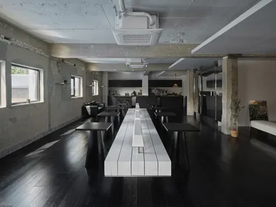 Empty modern restaurant with a long white table and black flooring, surrounded by chairs and well-lit by natural and artificial light